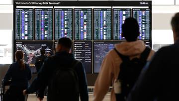 Tablica informacyjna na lotnisku z listą odlotów i przylotów, przed którą stoją pasażerowie.