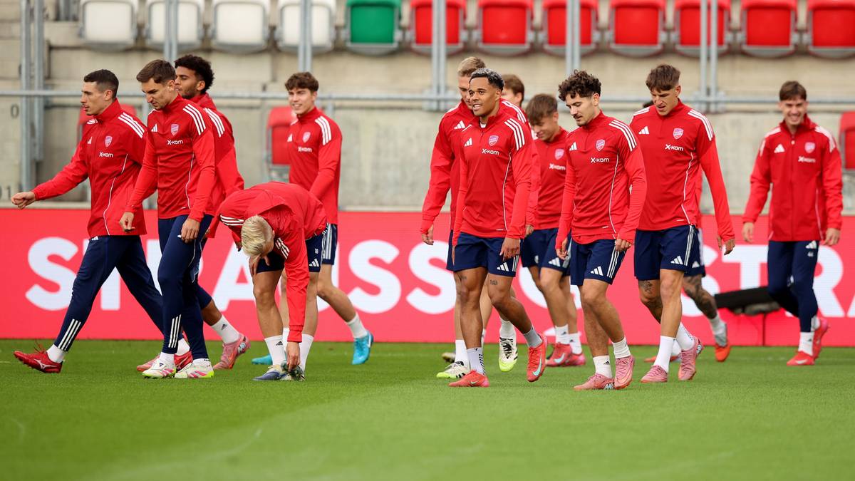 Grupa piłkarzy w czerwonych strojach treningowych na boisku, stojący obok siebie, z kilkoma zawodnikami na pierwszym planie, a w tle trybuny stadionu.