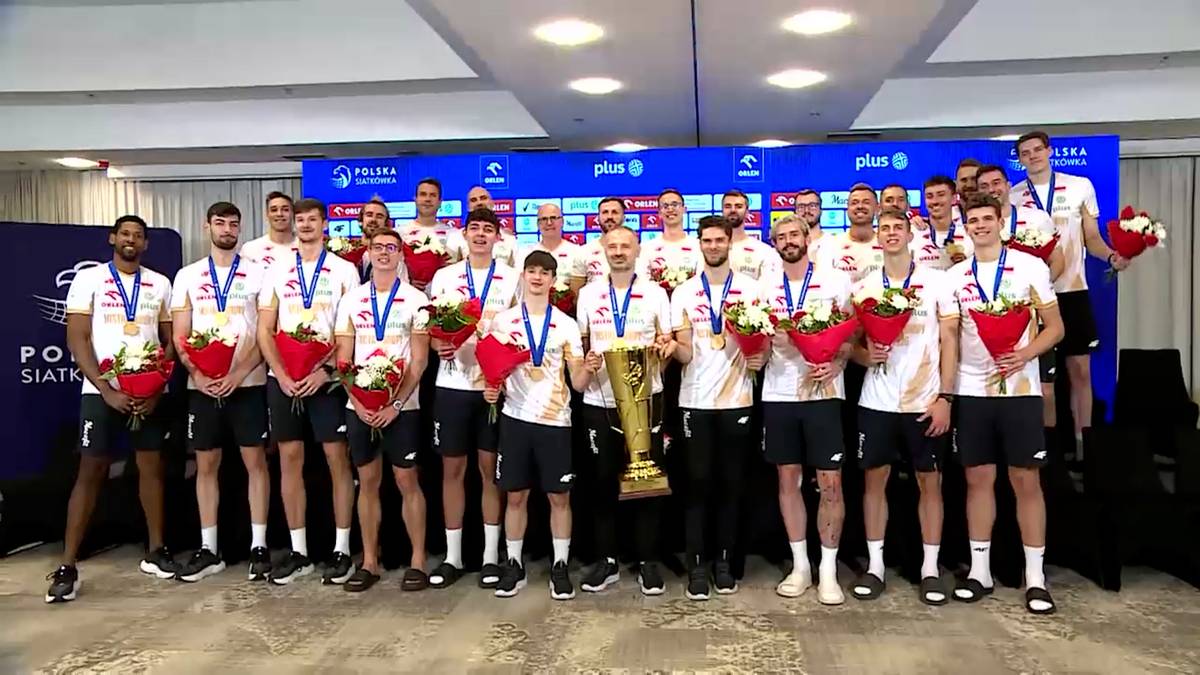 Grupa męskich siatkarzy z medalami i bukietami kwiatów pozuje z dużym złotym pucharem na tle niebieskiej ścianki sponsorskiej.