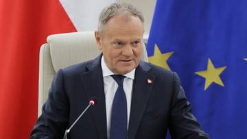 Donald Tusk, ubrany w ciemny garnitur i niebieski krawat, siedzi przy stole, przed nim mikrofon i dokumenty. W tle widoczne są flagi Polski i Unii Europejskiej.