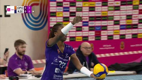 Zawodniczka w fioletowej koszulce z numerem 27 i piłką do siatk&oacute;wki w ręku przygotowuje się do zagrywki. W tle widoczni są inni ludzie, w tym kibice i sędziowie.