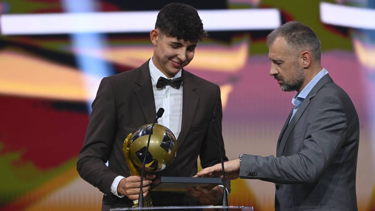 Młody mężczyzna w garniturze z muszką otrzymuje złotą statuetkę w kształcie piłki nożnej od starszego mężczyzny.