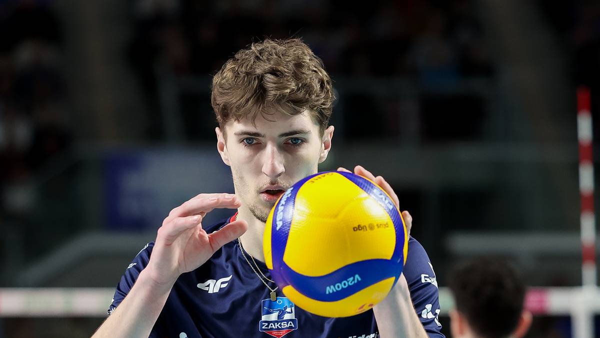 Siatkarz w niebieskiej koszulce z numerem 7 trzyma piłkę do siatk&oacute;wki przed sobą, przygotowując się do zagrywki. W tle widoczni są inni zawodnicy i trybuny.