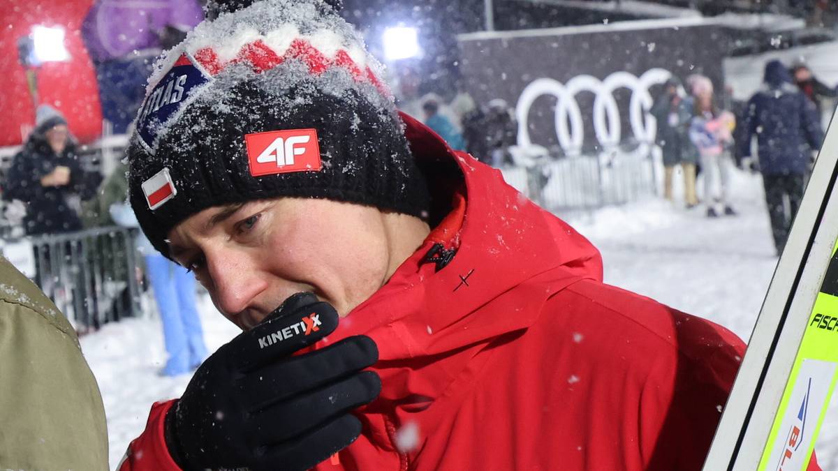 Kamil Stoch w czapce i kurtce narciarskiej, z rękawiczką na ustach, zwraca się do kibiców podczas zawodów w Zakopanem.