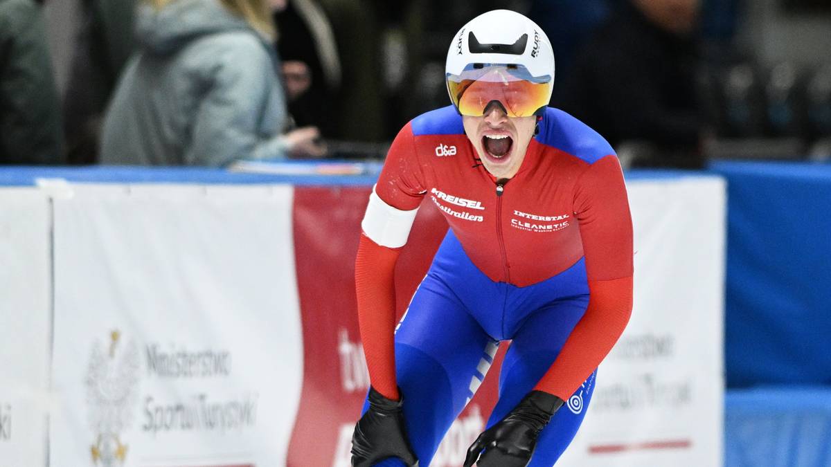 Panczenista w czerwono-niebieskim stroju z białym kaskiem i goglami na twarzy, z otwartymi ustami i okrzykiem radości na lodowisku.