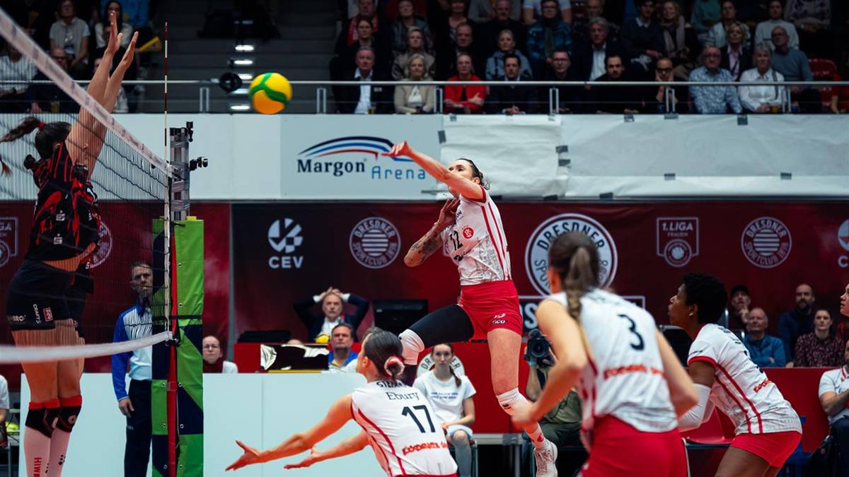 Siatkarka w białej koszulce z numerem 17 skacze do ataku, próbując przebić piłkę nad siatką, podczas gdy inne zawodniczki w czerwonych strojach po drugiej stronie boiska próbują ją zablokować.