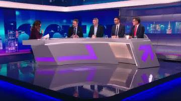 Cztery osoby w garniturach siedzą przy stole w studio telewizyjnym, rozmawiając z kobietą prowadzącą program. W tle widać nowoczesne oświetlenie i ekrany.