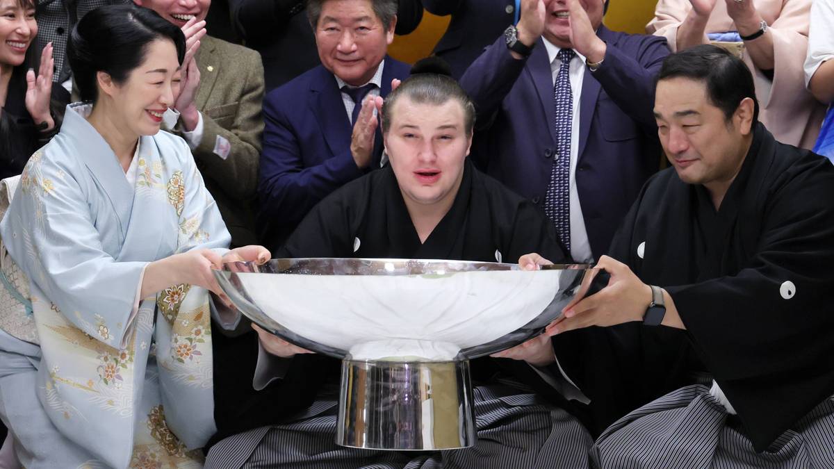 Młody mężczyzna w stroju sumo z wielką misą, otoczony przez ludzi w tradycyjnych strojach i tłum kibiców.