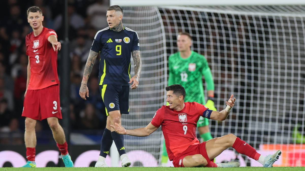 Piłkarze w akcji na boisku piłkarskim, jeden z nich leży na ziemi.