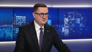 Mężczyzna w okularach i garniturze siedzi przed ekranem z nocnym widokiem miasta.