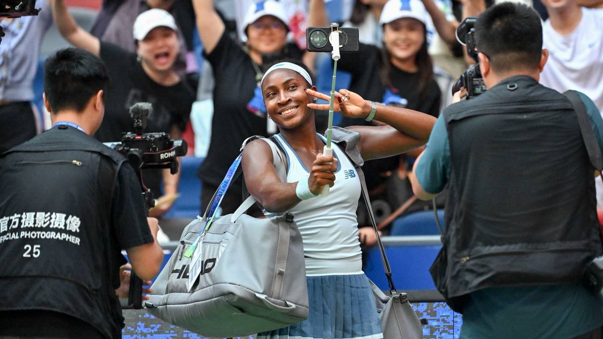 Uśmiechnięta tenisistka w stroju sportowym pozuje do zdjęcia z telefonem kom&oacute;rkowym w ręku, pokazując gest zwycięstwa palcami, podczas turnieju WTA w Wuhan. Wok&oacute;ł niej znajdują się kibice i dziennikarze z kamerami.