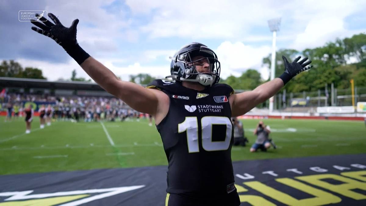 Gracz futbolu amerykańskiego w kasku i kamizelce z numerem 10 na plecach, z rękami uniesionymi do góry w geście zwycięstwa, na tle murawy i trybun stadionu.