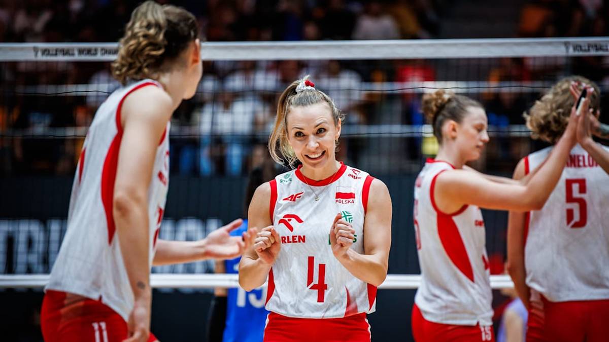 Uśmiechnięta siatkarka z numerem 4 na koszulce w białej bluzce z czerwonymi elementami, otoczona przez koleżanki z drużyny.