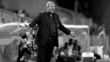 Trener piłkarski w garniturze, gestykulujący na boisku.