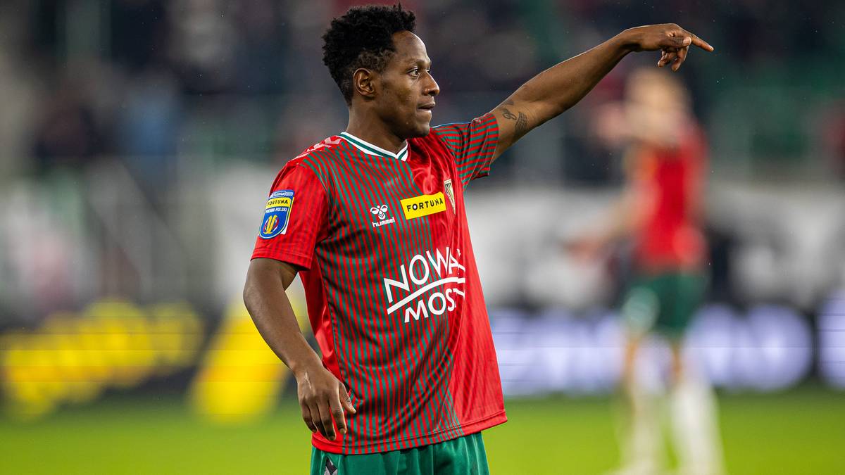 Piłkarz w czerwono-zielonej koszulce wskazuje palcem na bok, podczas meczu.