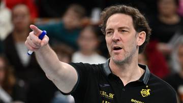 Trener siatk&oacute;wki w czarnej koszulce z emblematami drużyny, wskazujący palcem w kierunku boiska.