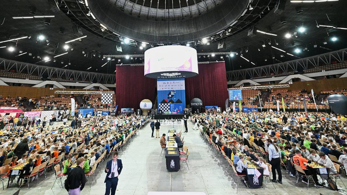 Szeroka sala widowiskowa z wieloma rzędami siedzeń zajętymi przez uczestników turnieju. Na środku areny ustawione są stoły, przy których siedzą zawodnicy. Na tle sceny znajduje się duży ekran.