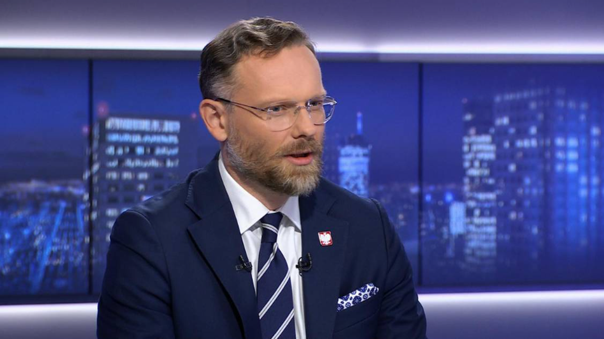 Mężczyzna w garniturze i okularach siedzi w studiu telewizyjnym.