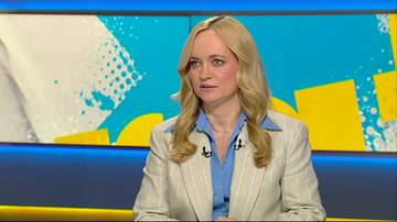 Kobieta z blond włosami, ubrana w jasną marynarkę w prążki i niebieską koszulę, siedzi i m&oacute;wi. Tło za nią jest niebieskie z ż&oacute;łtymi i białymi geometrycznymi kształtami.