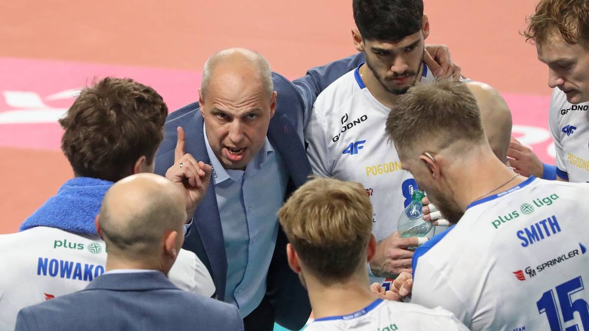 Trener siatkarzy w niebieskiej koszuli zwraca się do graczy w białych strojach, wskazując palcem.