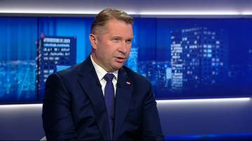 Mężczyzna w granatowym garniturze z białą koszulą i granatowym krawatem przemawia w studiu telewizyjnym. Tło stanowi niebieska ściana z grafiką przedstawiającą budynki nocnego miasta.