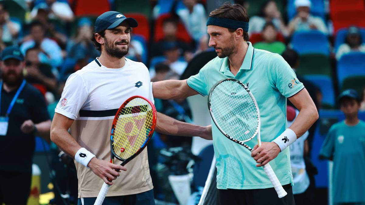 Dwóch tenisistów, jeden w białej koszulce z niebieskimi pasami, drugi w turkusowej koszulce, stoi na korcie tenisowym, trzymając rakiety i patrząc na siebie.