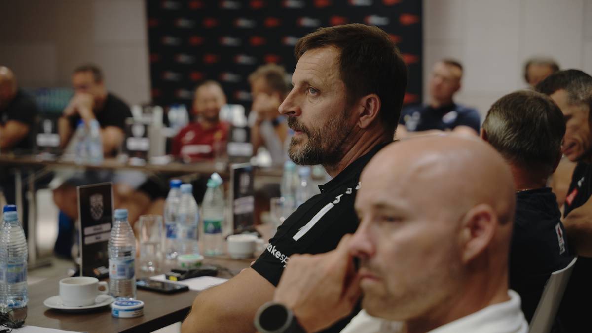 Zbliżenie na twarz mężczyzny w średnim wieku o kr&oacute;tkich, ciemnych włosach i zaroście, siedzącego przy stole. Mężczyzna jest ubrany w czarną koszulkę z białym pasem i patrzy w bok. W tle widać innych ludzi siedzących przy stołach.