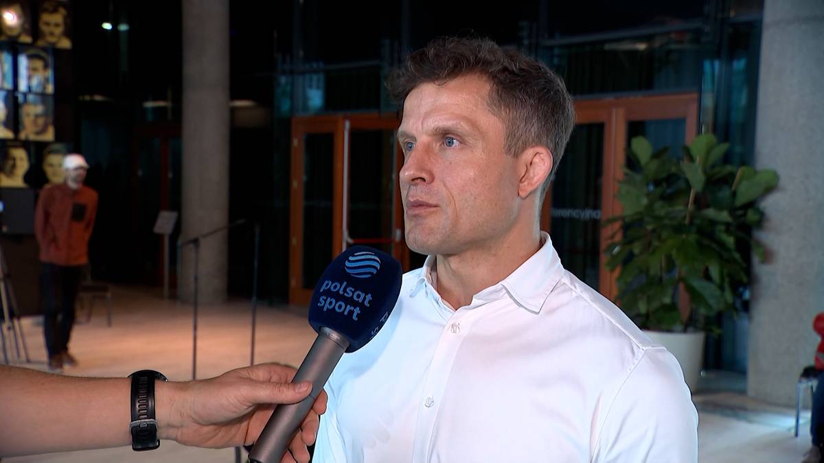 Mężczyzna w białej koszuli z bliska, trzymający się z dala od mikrofonu z logo Polsat Sport, który jest trzymany przez niewidzialną osobę. W tle znajduje się kilka osób i roślin.