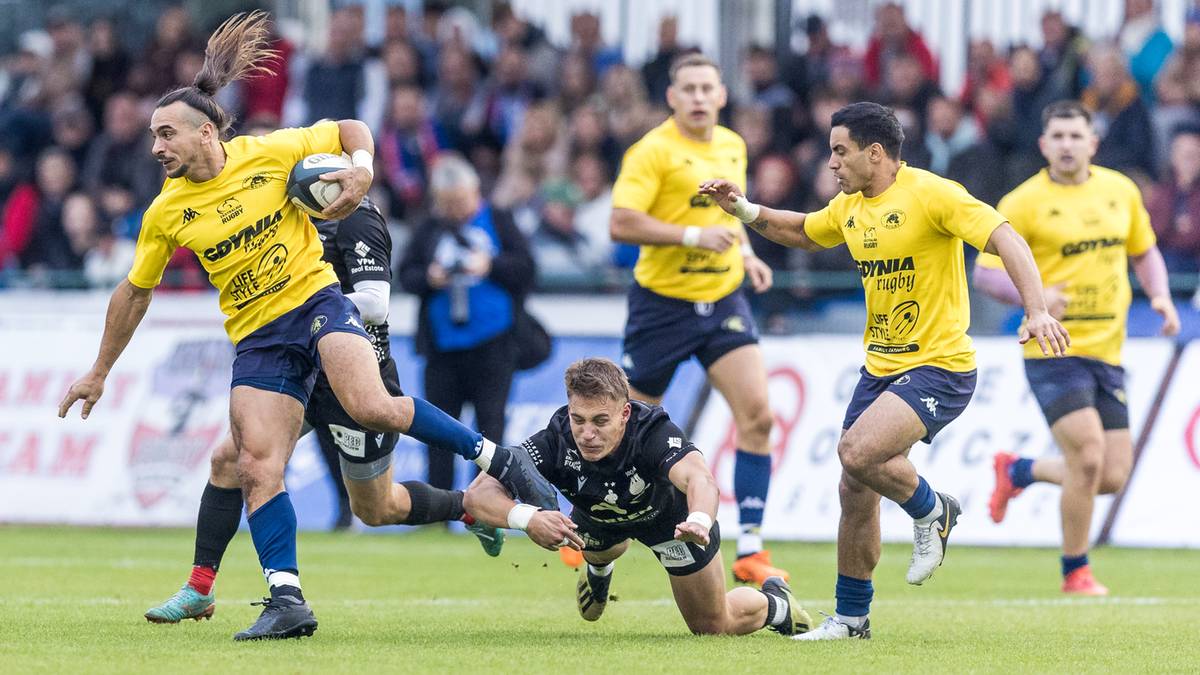 Zawodnicy drużyny rugby w akcji podczas meczu, jeden z nich pr&oacute;buje odebrać piłkę rywalowi, kt&oacute;ry jest w posiadaniu futbol&oacute;wki.