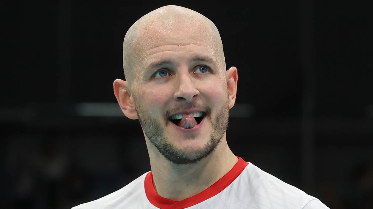 Siatkarz Bartosz Kurek w białej koszulce reprezentacji Polski z czerwonymi wstawkami, ogolona głowa, pokazuje język.