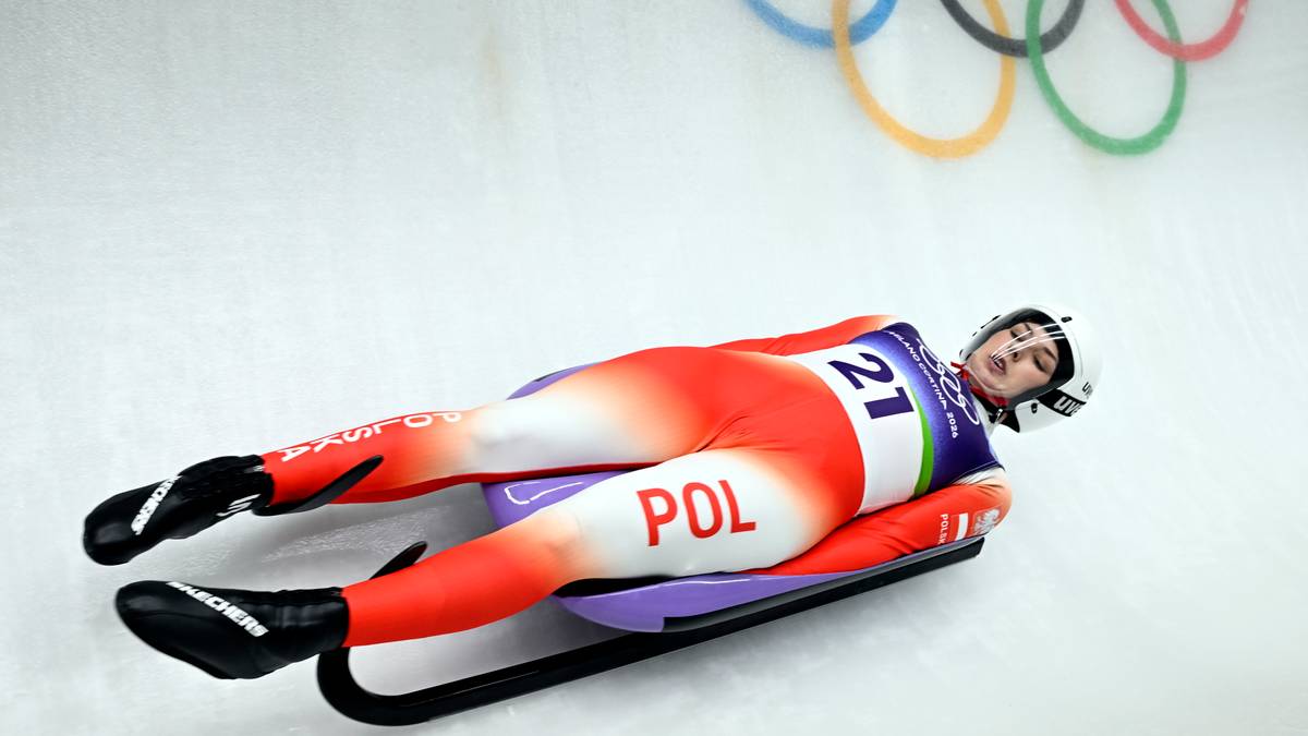 Zawodniczka saneczkarstwa na torze podczas zawod&oacute;w, ubrana w czerwono-biały kombinezon z napisem "POL" na nogawce. W tle widoczne k&oacute;łka olimpijskie.