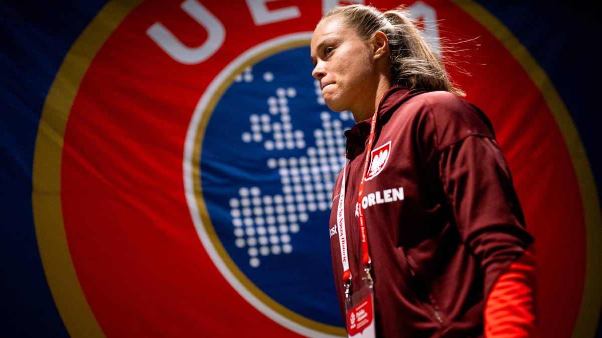 Piłkarka reprezentacji Polski w bordowej bluzie treningowej, z widocznym herbem Polski na piersi, na tle okrągłego logo UEFA z mapą Europy.