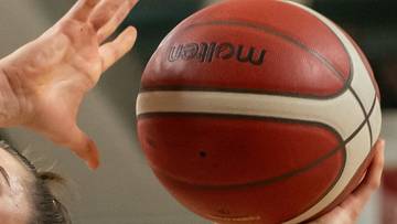 Koszykarka w białej koszulce z numerem 73 i napisem Enea rzuca do kosza, podczas gdy zawodniczka z numerem 95 w zielonej koszulce pr&oacute;buje ją zablokować.