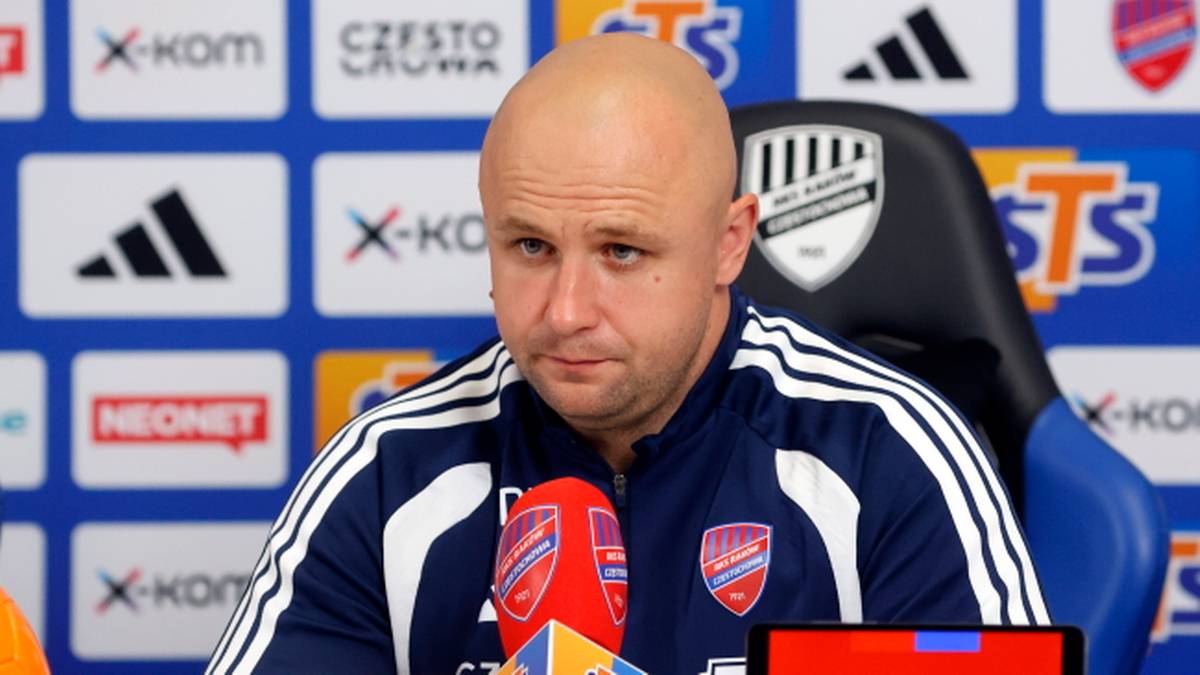 Mężczyzna w kurtce sportowej siedzi przed ścianą z logo sponsorów, patrząc na mikrofony.