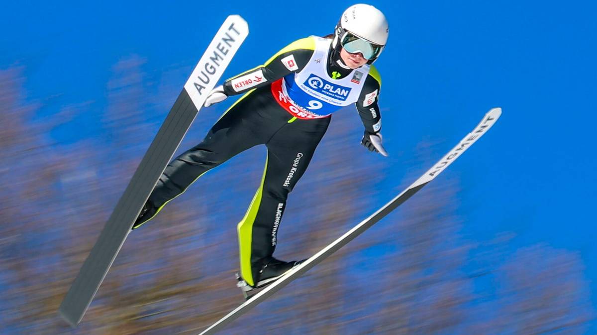 Zawodniczka w stroju narciarskim i kasku leci w powietrzu na nartach podczas skoku narciarskiego.