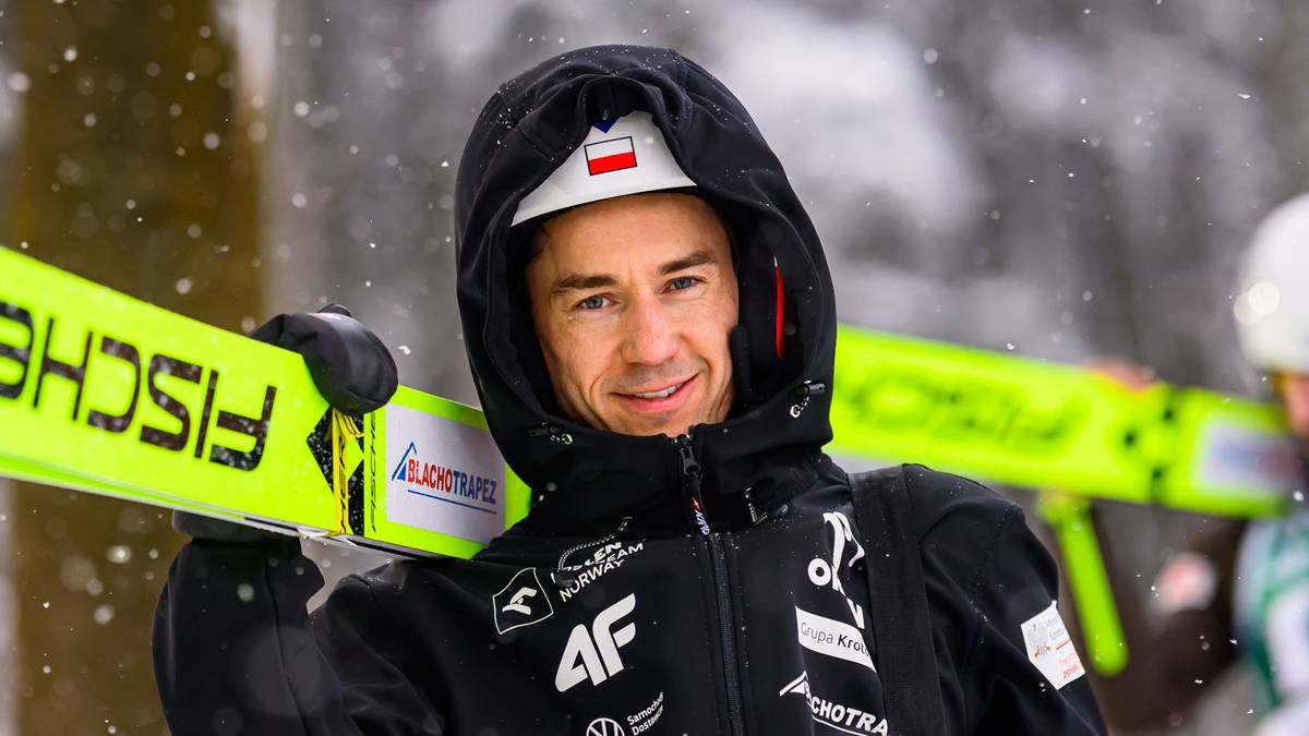 Sportowiec w zimowej kurtce i kapturze trzyma narty biegowe na ramieniu.