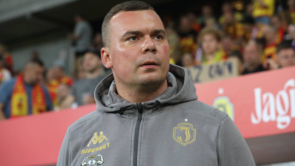 Mężczyzna w szarej bluzie z kapturem i logotypami, patrzy w bok.