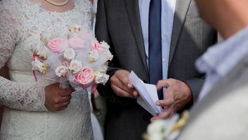 Zbliżenie na pannę młodą w białej sukni ślubnej trzymającą bukiet r&oacute;żowych kwiat&oacute;w oraz pana młodego w garniturze trzymającego kartkę papieru.