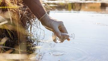 Dłoń w rękawiczce pobiera pr&oacute;bkę wody.