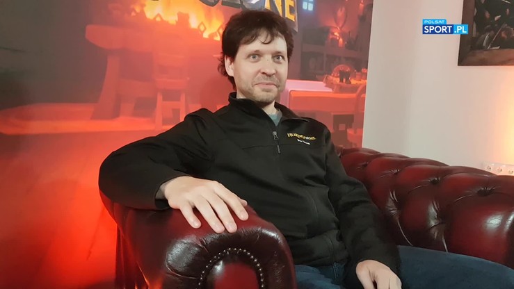 Mężczyzna w czarnej kurtce z logo Hearthstone siedzi na czerwonej skórzanej kanapie.