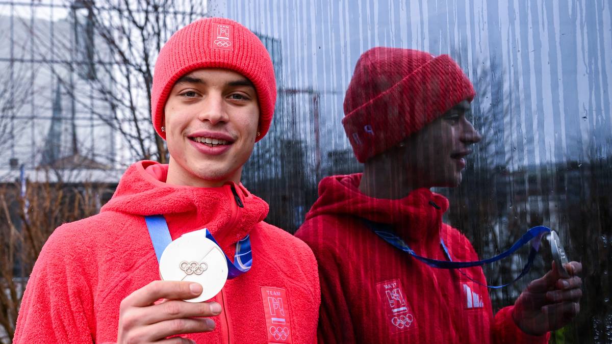 Młody mężczyzna w czerwonym stroju sportowym trzyma medal olimpijski na szyi, z uśmiechem prezentuje go przed obiektywem.