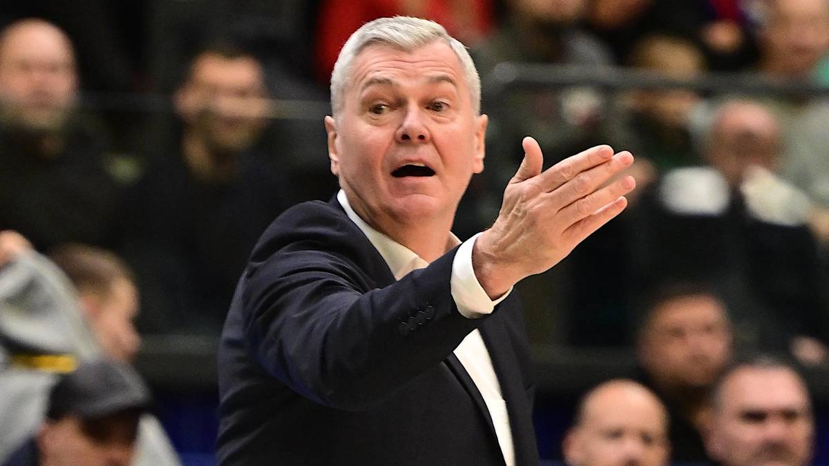 Trener w garniturze gestykuluje ręką w stronę boiska.