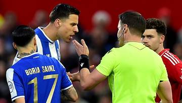 Sędzia w neonowo-zielonej koszulce rozmawia z dwoma piłkarzami w niebieskich i czerwonych strojach.