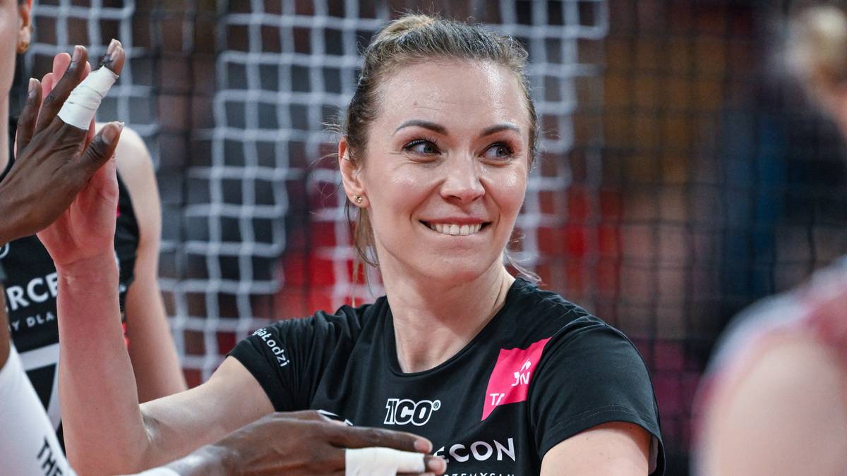 Uśmiechnięta siatkarka w czarnej koszulce z różowym elementem, na tle siatki i innych zawodniczek.