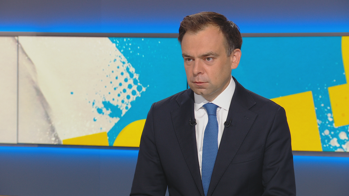 Mężczyzna w garniturze i krawacie siedzi przed ekranem z niebieskimi i żółtymi wzorami.