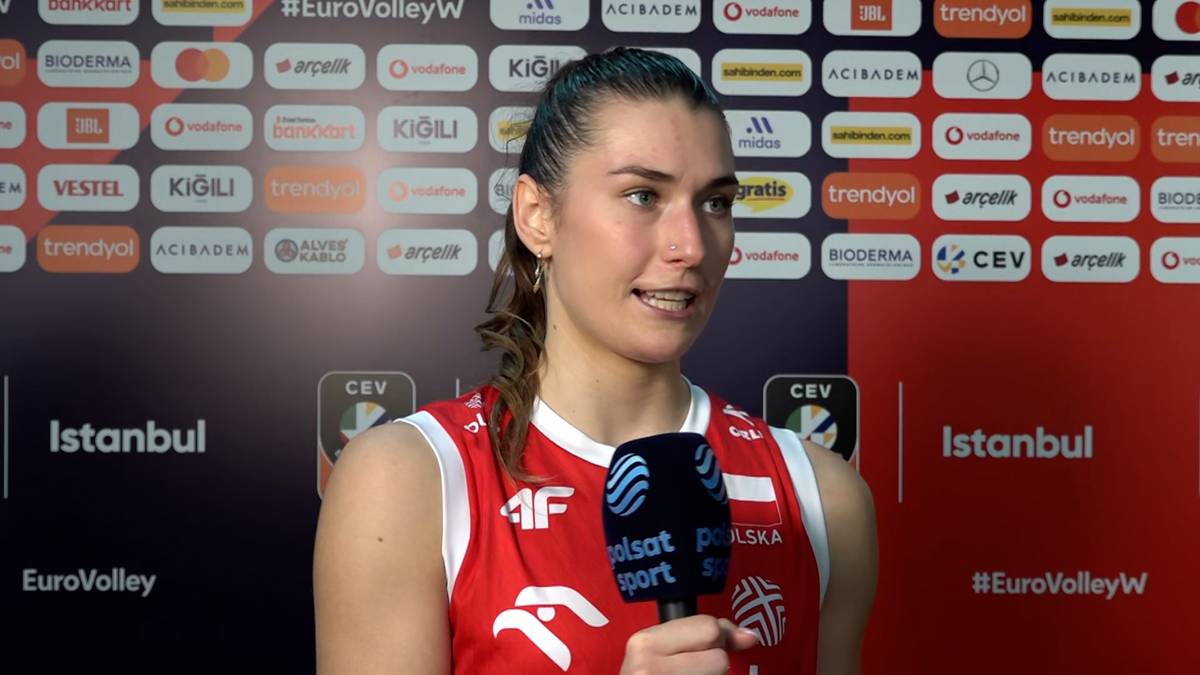 Portret kobiety w stroju sportowym z mikrofonem w ręku, udzielającej wywiadu na tle logotypów sponsorów.