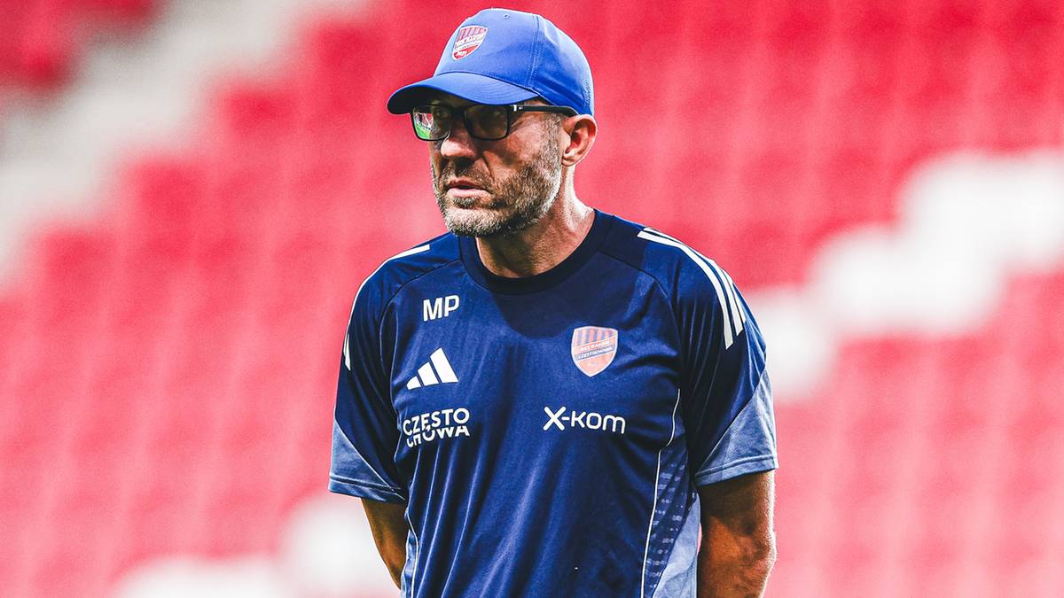 Trener piłkarski w niebieskiej czapce i okularach na tle stadionu z czerwonymi siedzeniami.