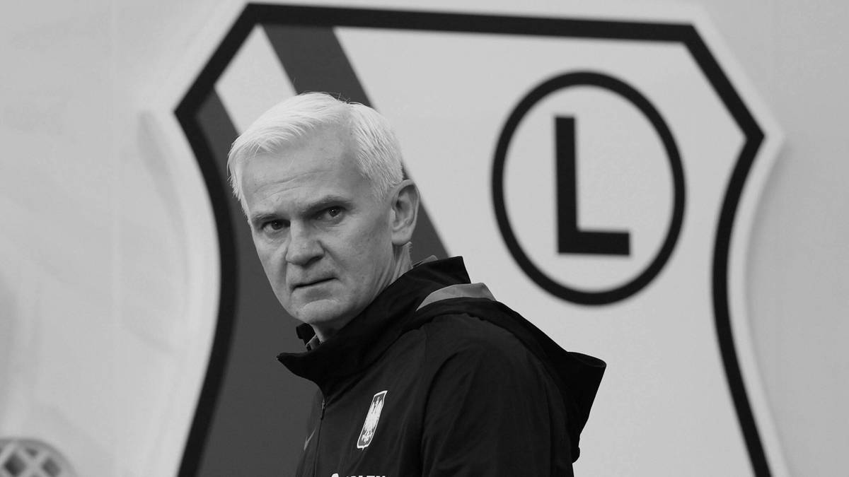 Jacek Magiera w czarno-białym zdjęciu, z herbem Legii Warszawa w tle.