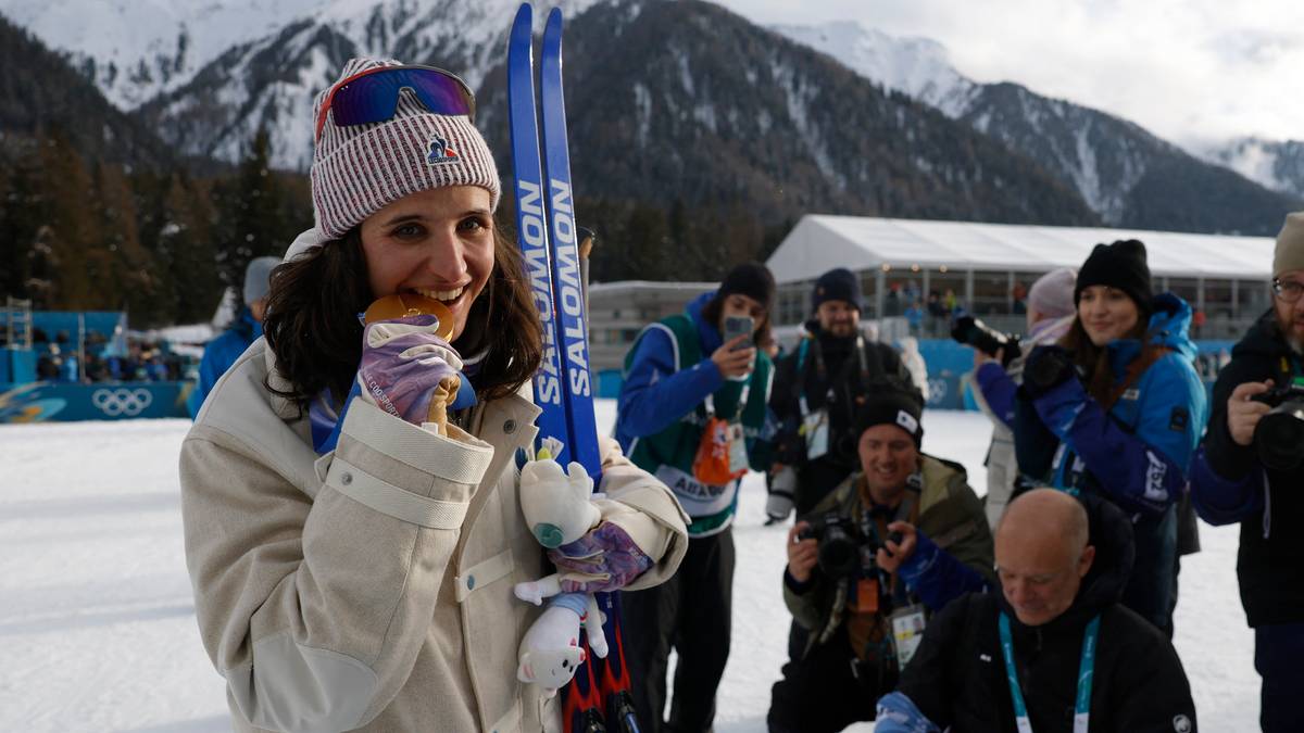 Biathlonistka Julia Simon z nartami trzyma w ustach kawałek jedzenia, uśmiecha się do obiektywu.