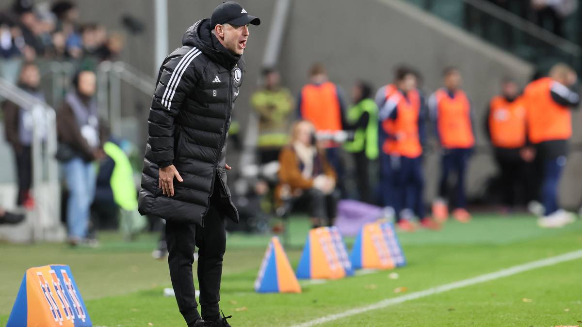 Trener piłkarski w czapce i długiej kurtce stoi na zielonej murawie, patrząc w bok.